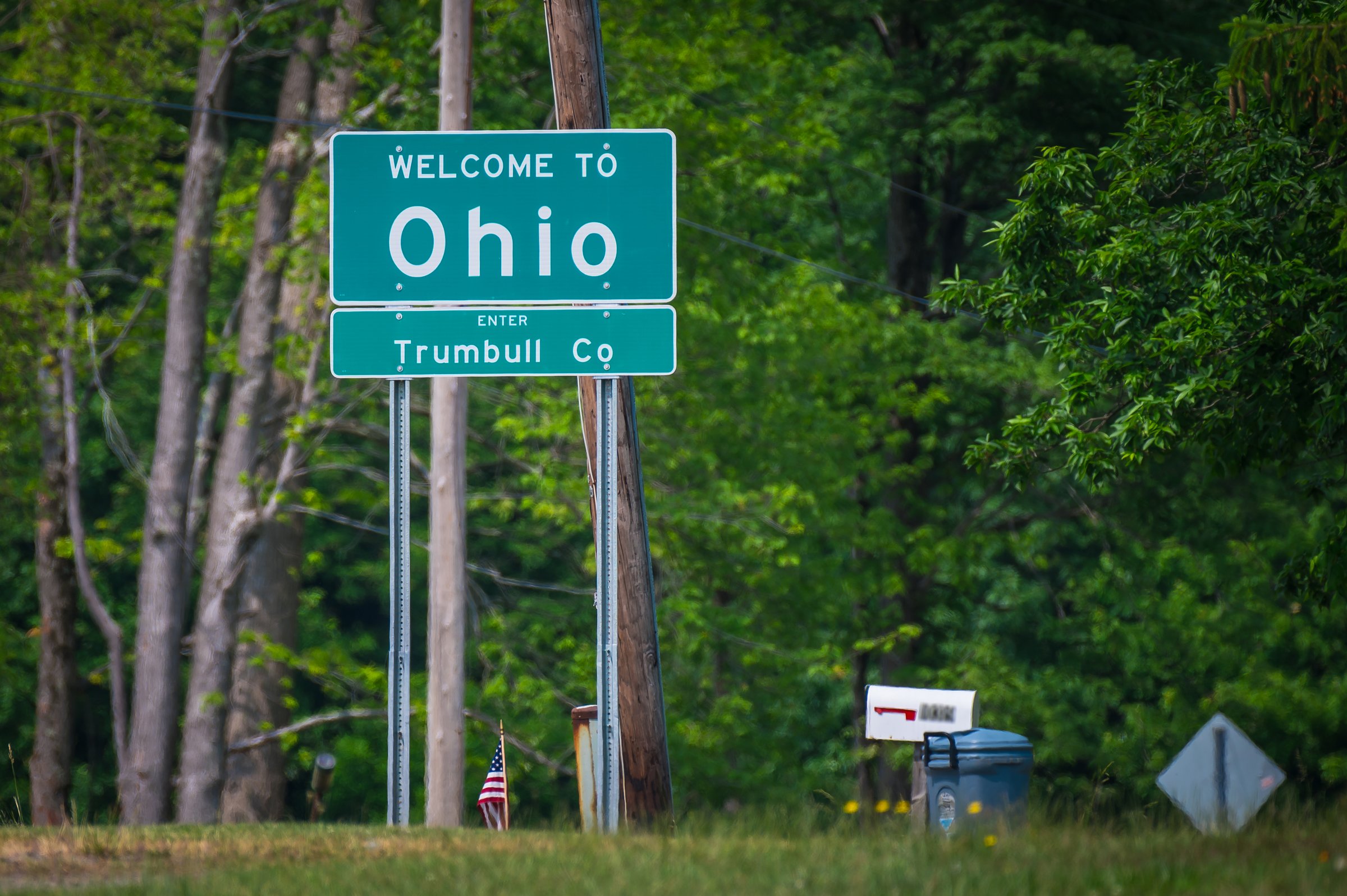 Green highway sign reading 'Welcome to Ohio, Enter Trumbull Co' in front of trees